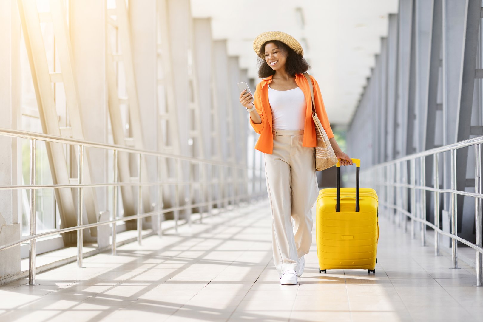 Stylish young black woman walking through airport with a yellow suitcase, smiling while looking at phone. Casual travel style with hat and tote bag, copy space