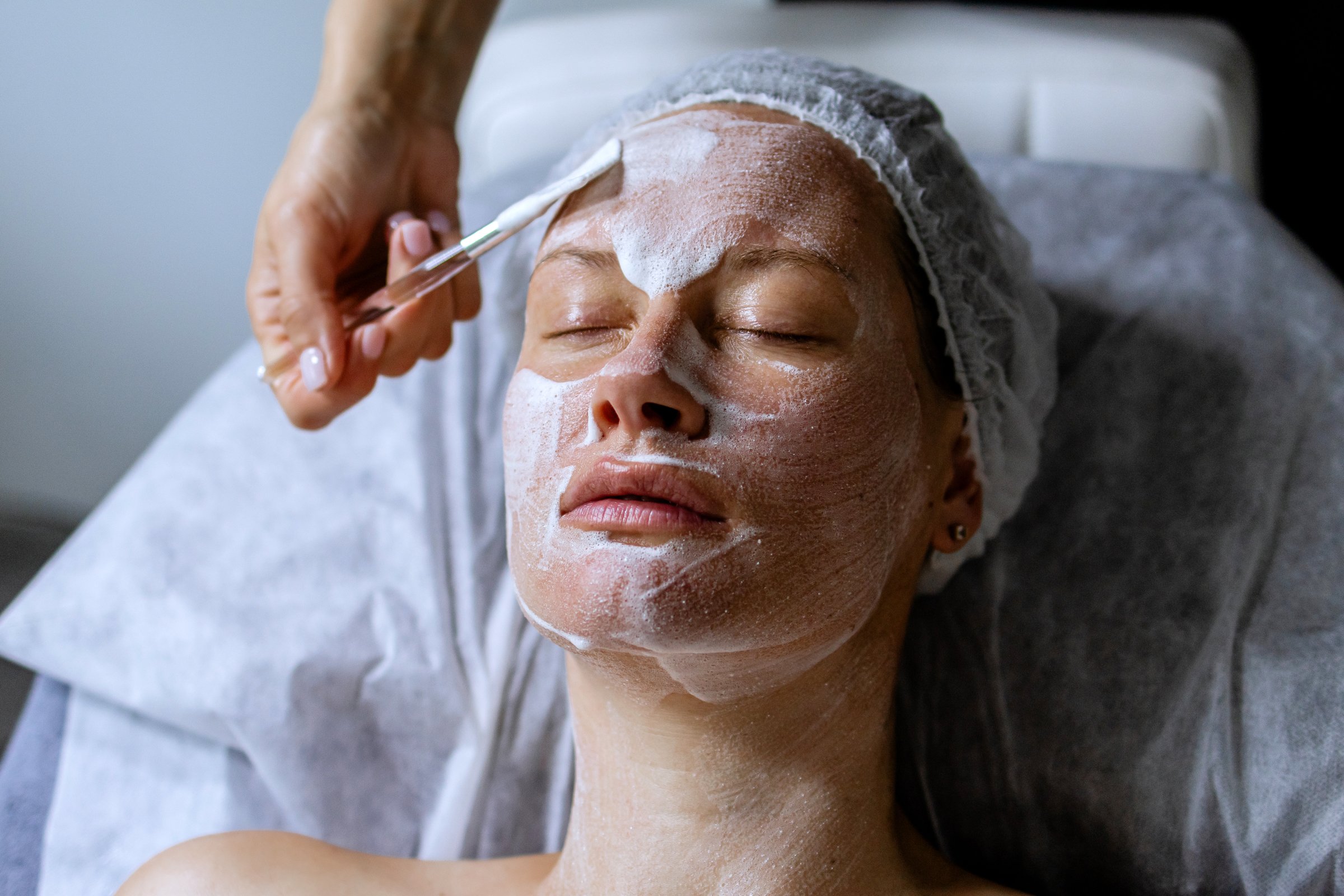 Middle aged female receiving facial anti aged massage in beauty salon. Skincare concept. Cosmetology specialist making facial lifting massage, appling cosmetic product to facial skin with brush