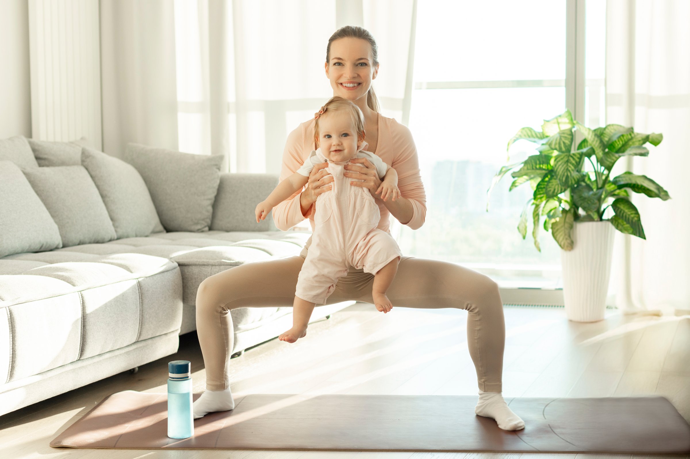 Happy European mother with little daughter on hands making squats at home, training on fitness mat, enjoying domestic workouts and motherhood