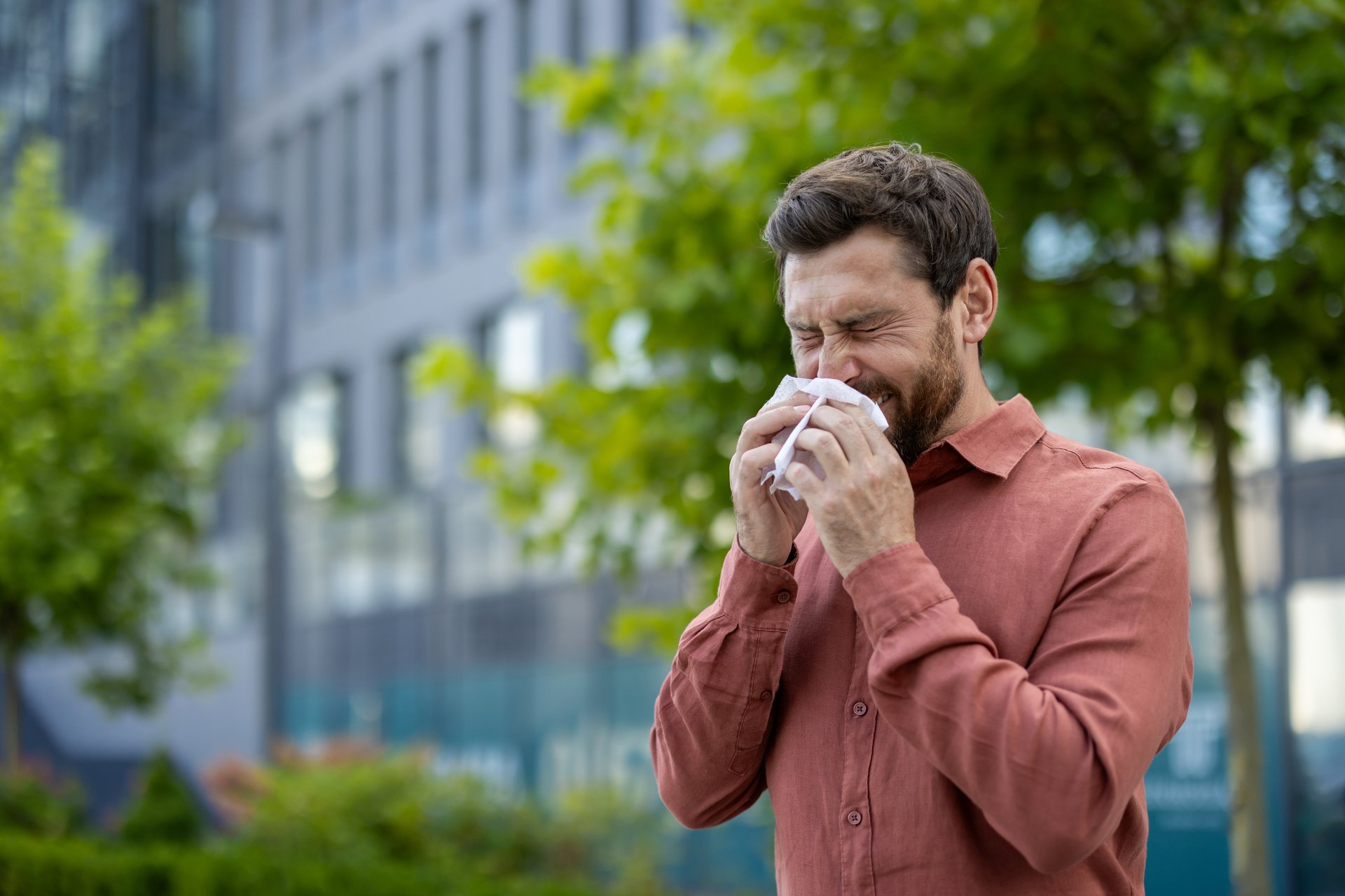 Mature man sneezes outside in urban landscape, reflecting discomfort due to allergies. Surrounded by modern buildings and greenery, capturing essence of city environment.