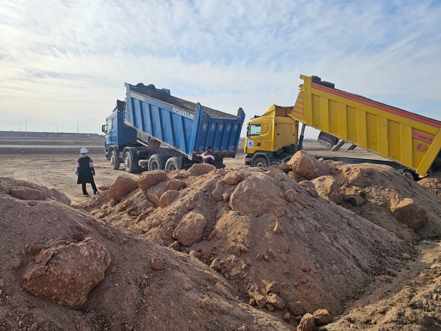 Two dump trucks unloading dirt at a construction site under a cloudy sky, with workers nearby and piles of rocks in the foreground.