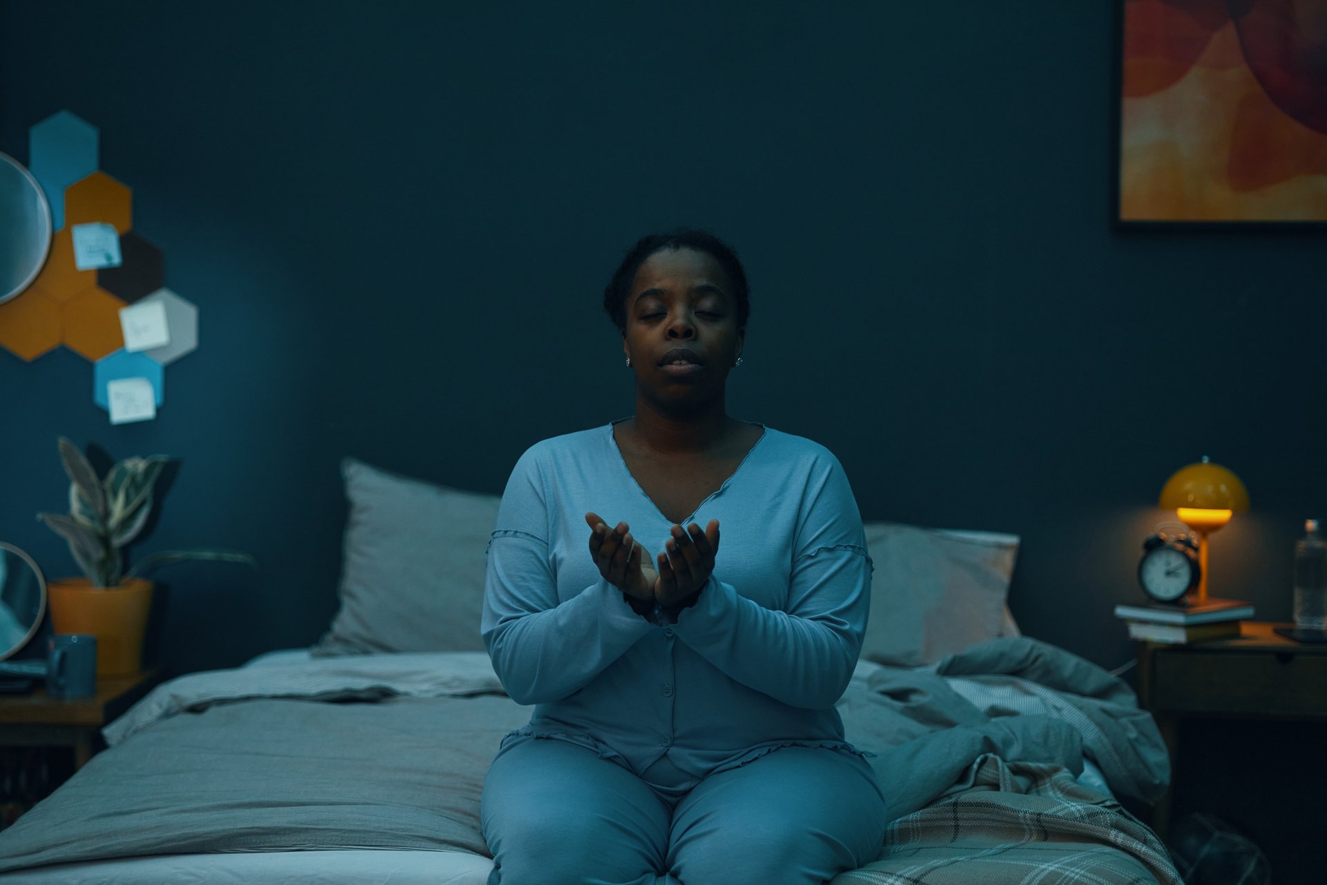 Woman sitting on bed in meditation pose in dimly lit room. Room decorated with hexagonal wall art, bedside table with a lamp, and books