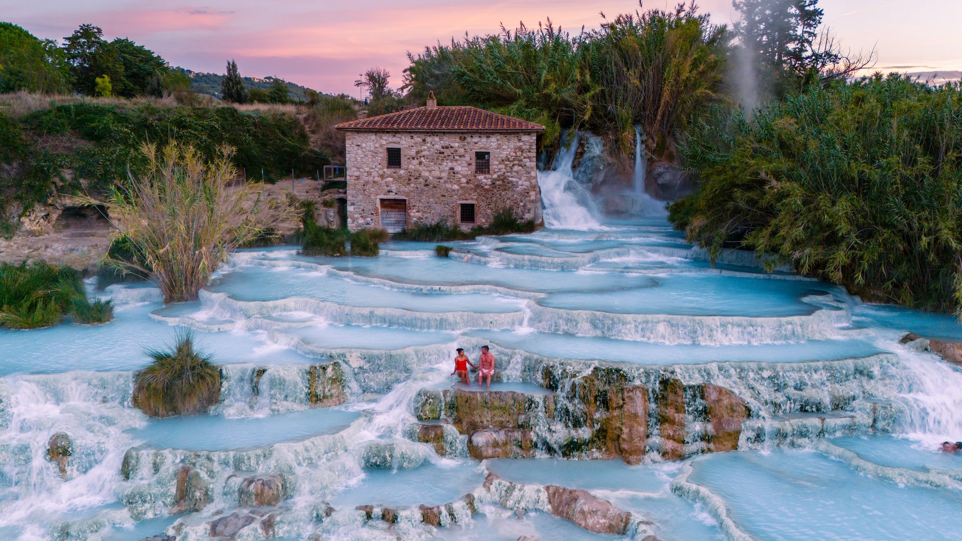 a couple enjoy the warm, mineral-rich waters of Saturnia's thermal baths, surrounded by lush greenery and historic stone buildings as the sun sets, creating a magical atmosphere in Italy
