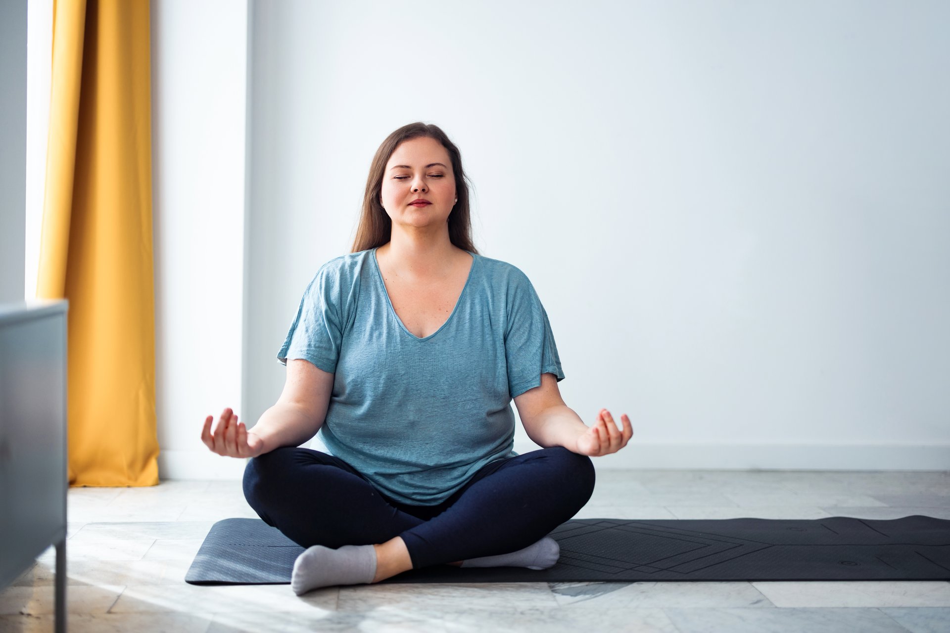 A woman practicing meditation while seated in a cross-legged pose on a yoga mat indoors, enjoying a sense of calmness and relaxation.