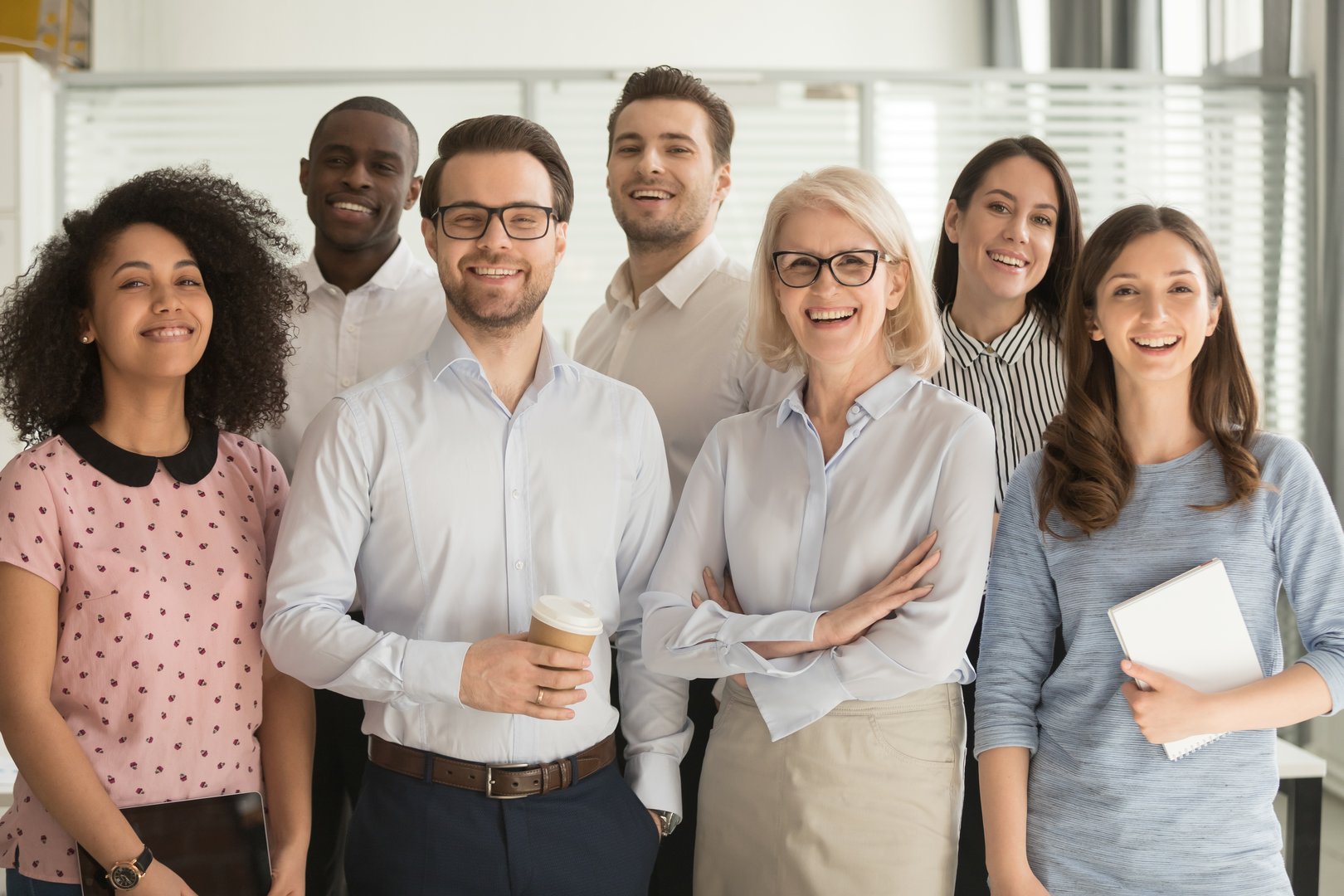 Smiling multiethnic employees standing looking at camera making team picture in office together, happy diverse work group or department laugh posing for photo at workplace, show unity and cooperation