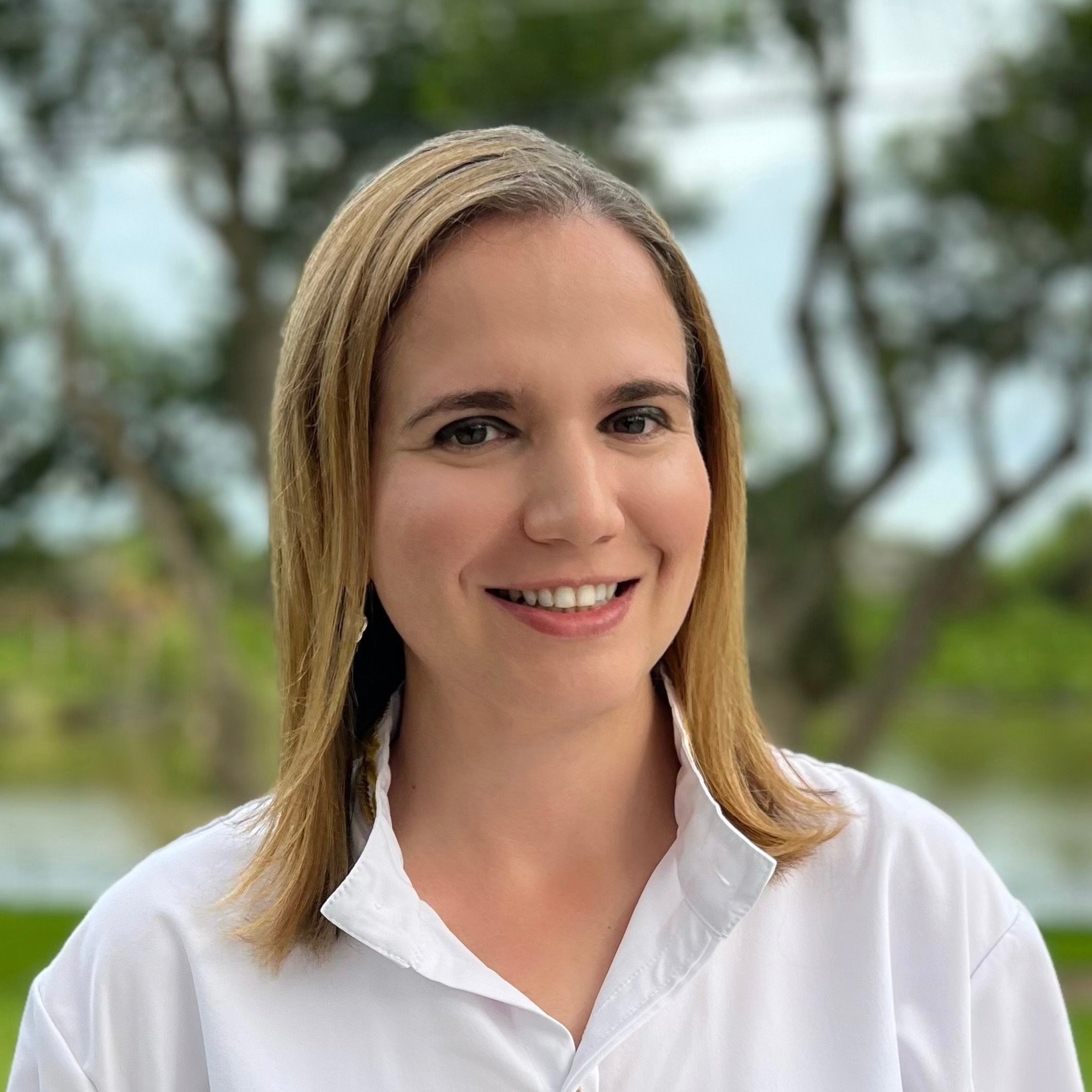 Smiling person with shoulder-length blonde hair wearing a white shirt, standing outdoors with trees in the background.