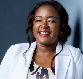 A woman in a white blazer, smiling, with dark hair and a patterned top. She stands against a plain background.