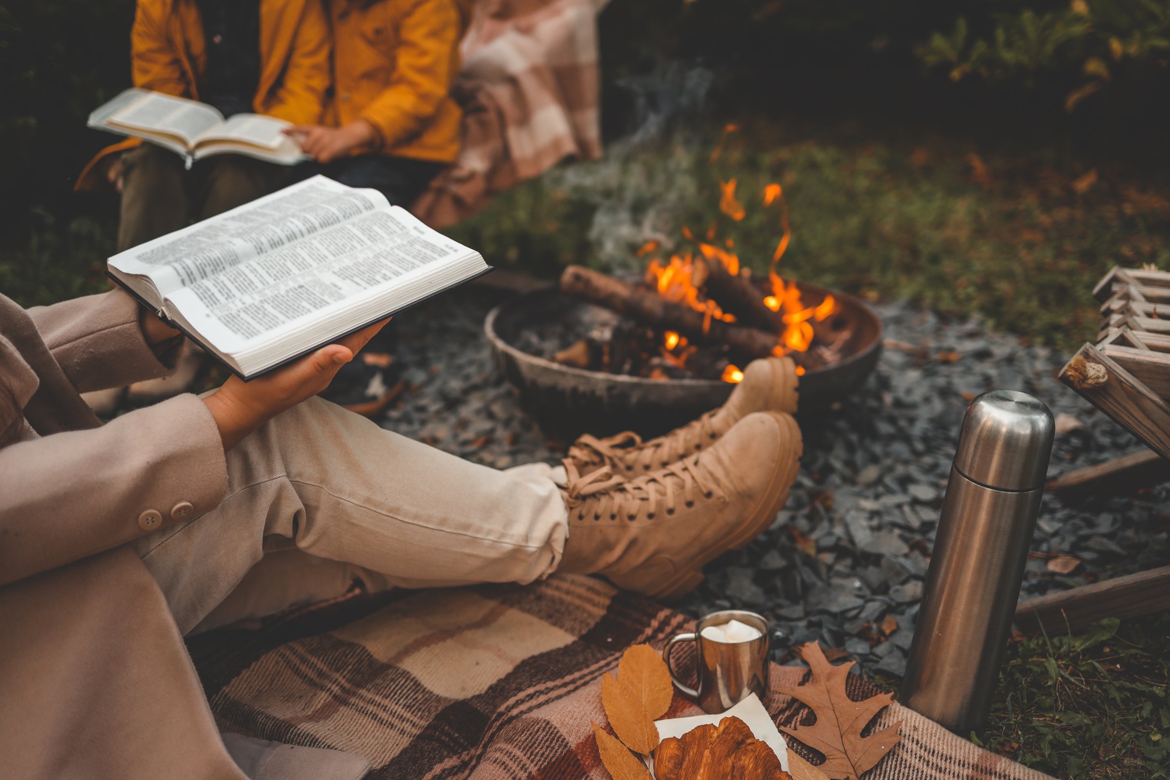 Reading the Bible by a warm campfire during autumn. A cozy scene with boots, blanket, hot drink with marshmallows, croissant, and colorful leaves.