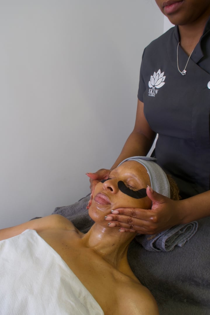 Person receiving a facial massage with eye patches in a spa setting, wearing a headband and covered in a white towel.