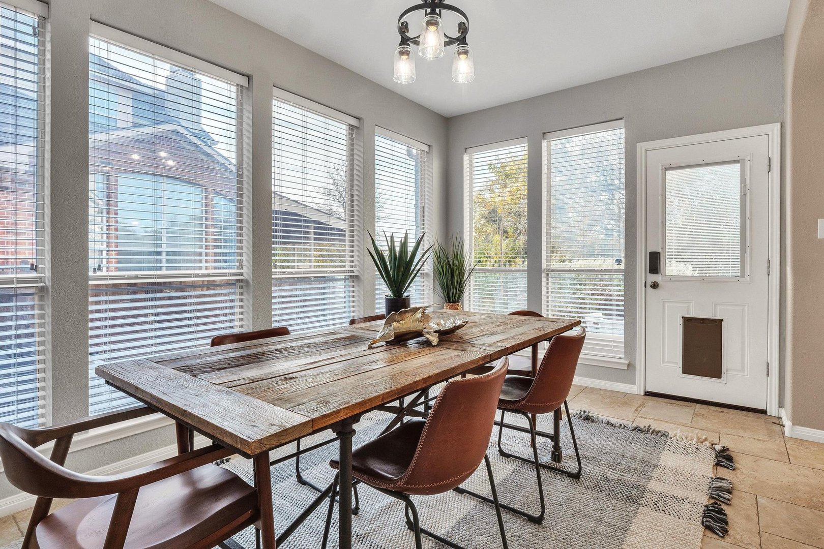 Dining area with wooden table, brown chairs, potted plant, and large windows letting in natural light.