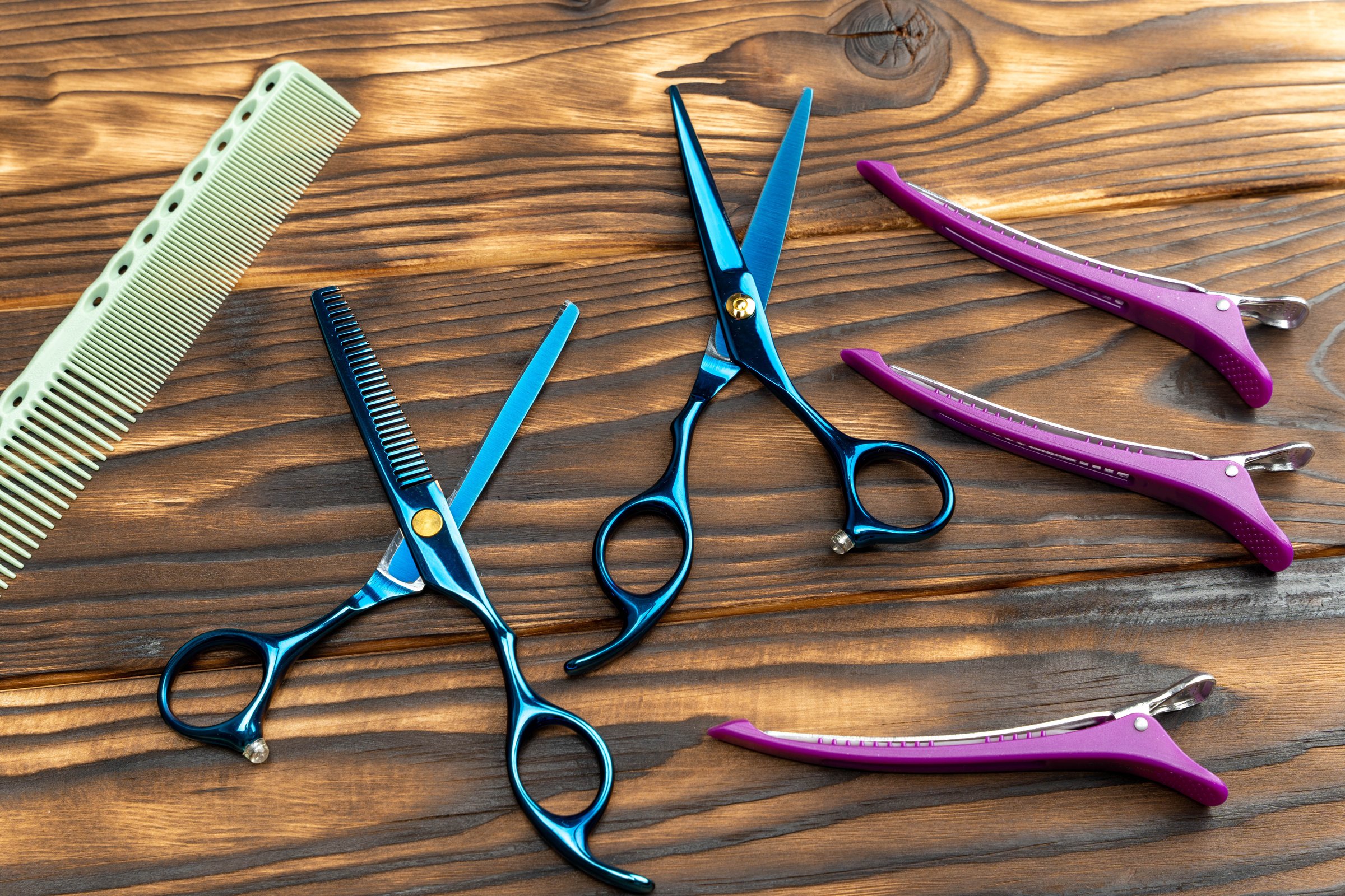 Hairdressing tools displayed on wooden surface, featuring blue scissors, pink clips, and green comb, highlighting essential equipment for professional hairstyling and grooming