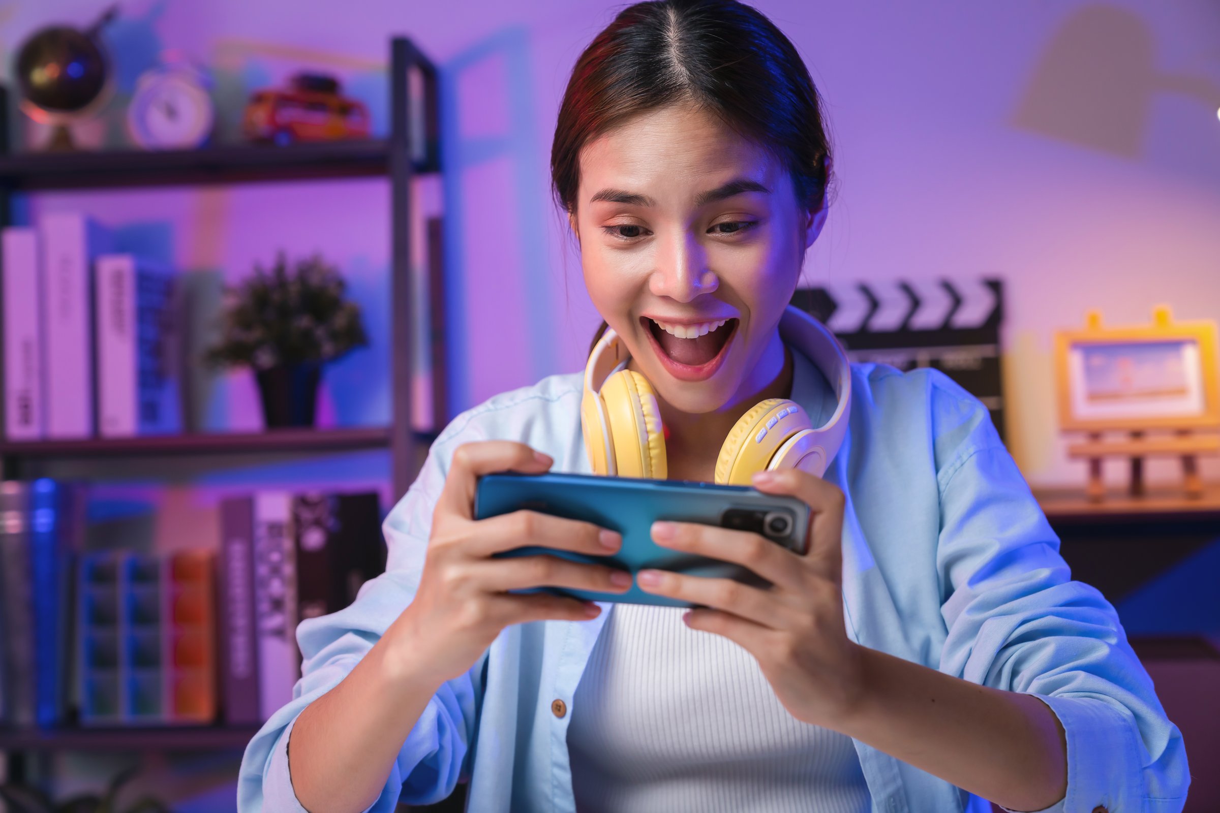Excited Young Asian woman wearing headset and playing online game on smartphone at night modern house.