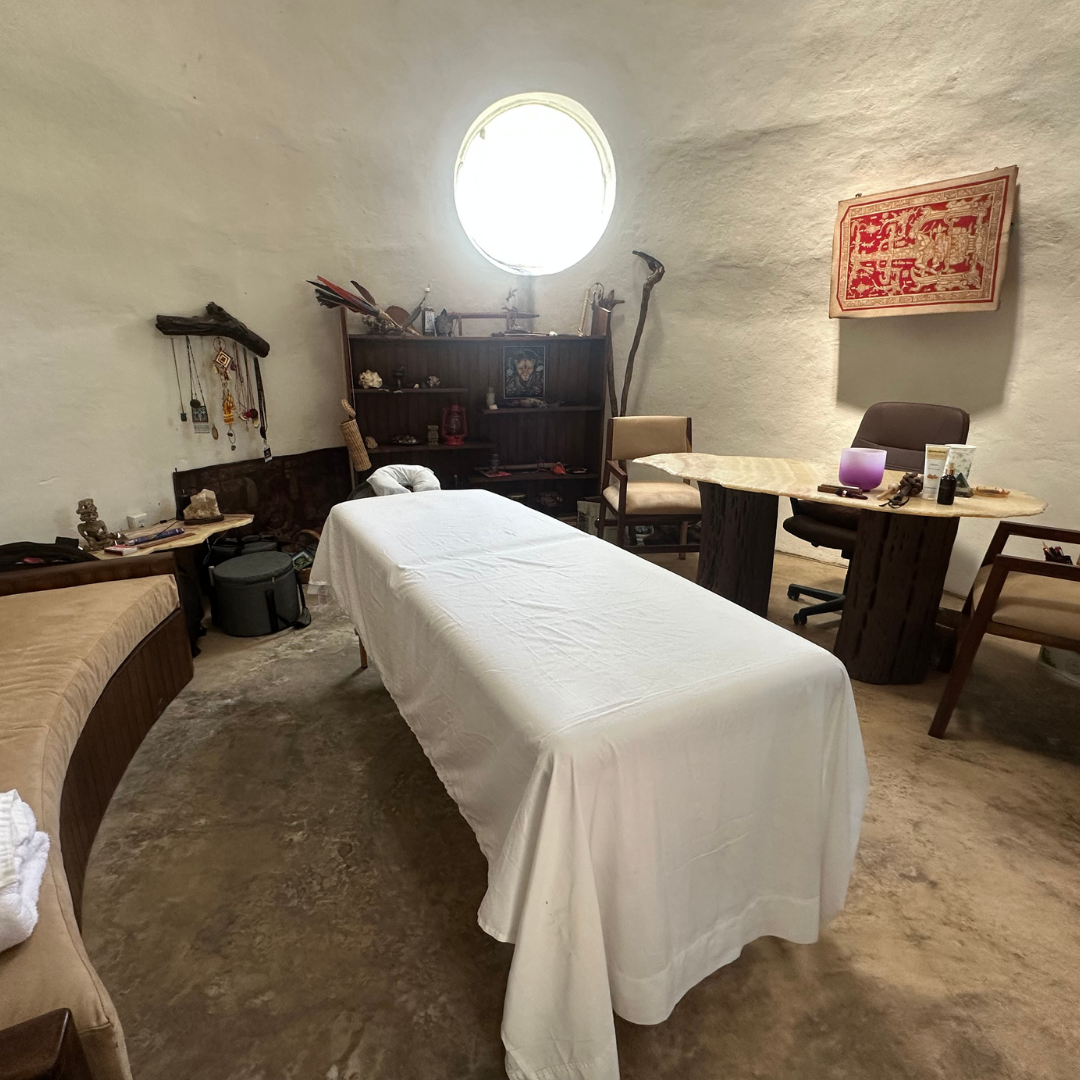 Cozy massage room with a massage table, circular window, artwork, chairs, and wooden shelves displaying various items.