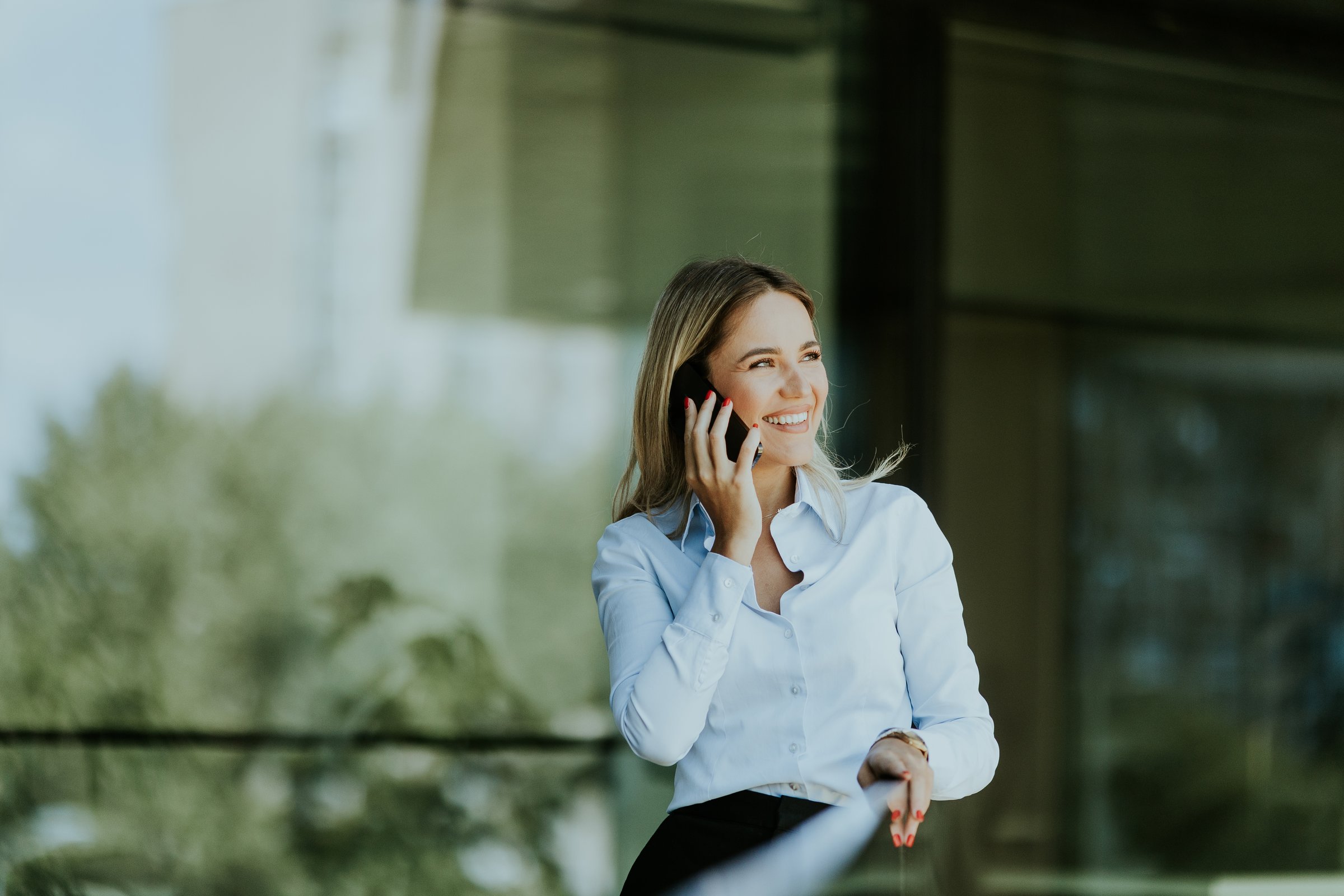 Businesswoman smiles while talking on her smartphone outside a modern office building on a sunny day, dressed in a white blouse and dark skirt