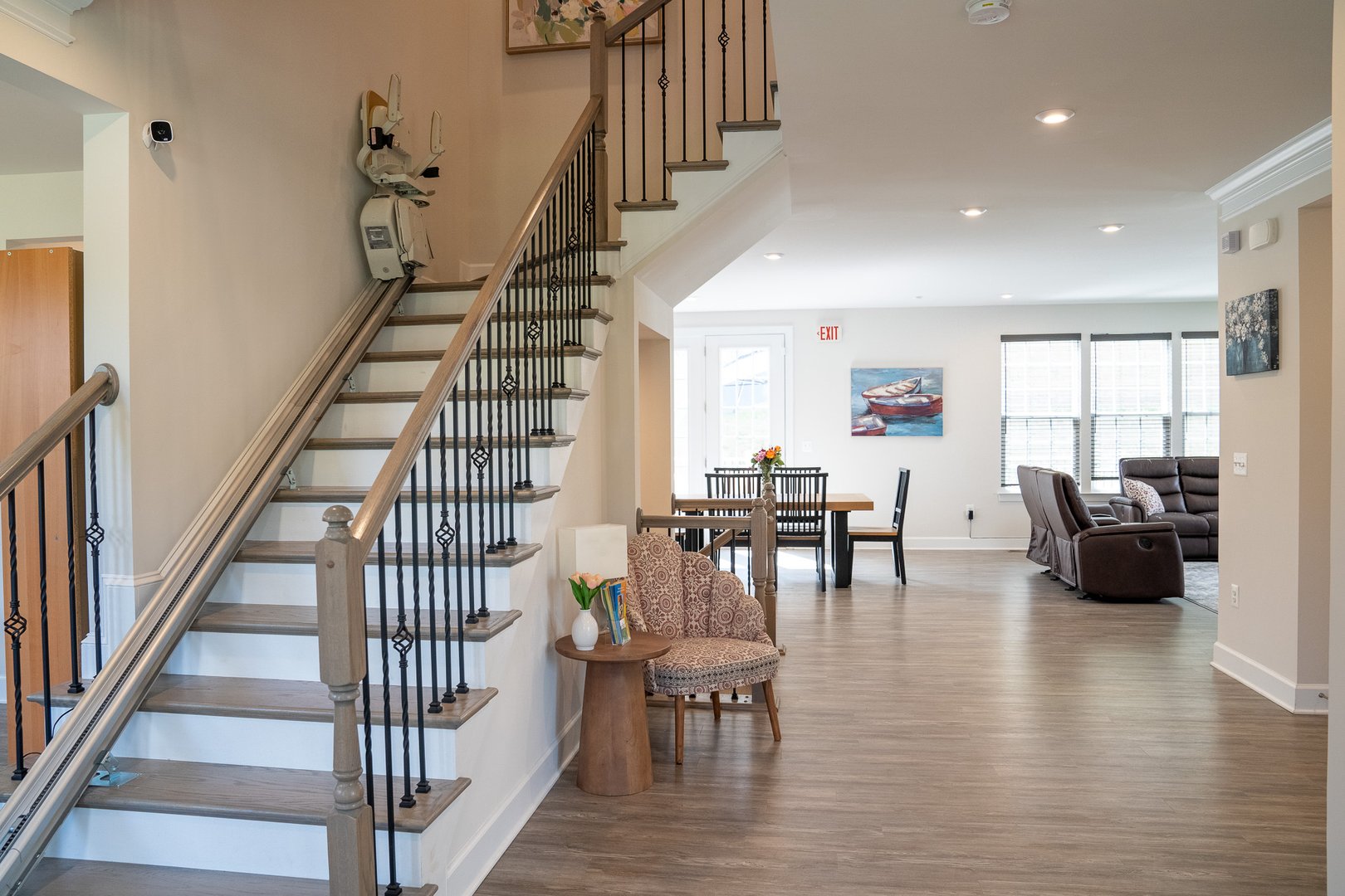 Cozy living space with a staircase, dining table, chairs, and brown sofa. Wooden flooring and light walls.