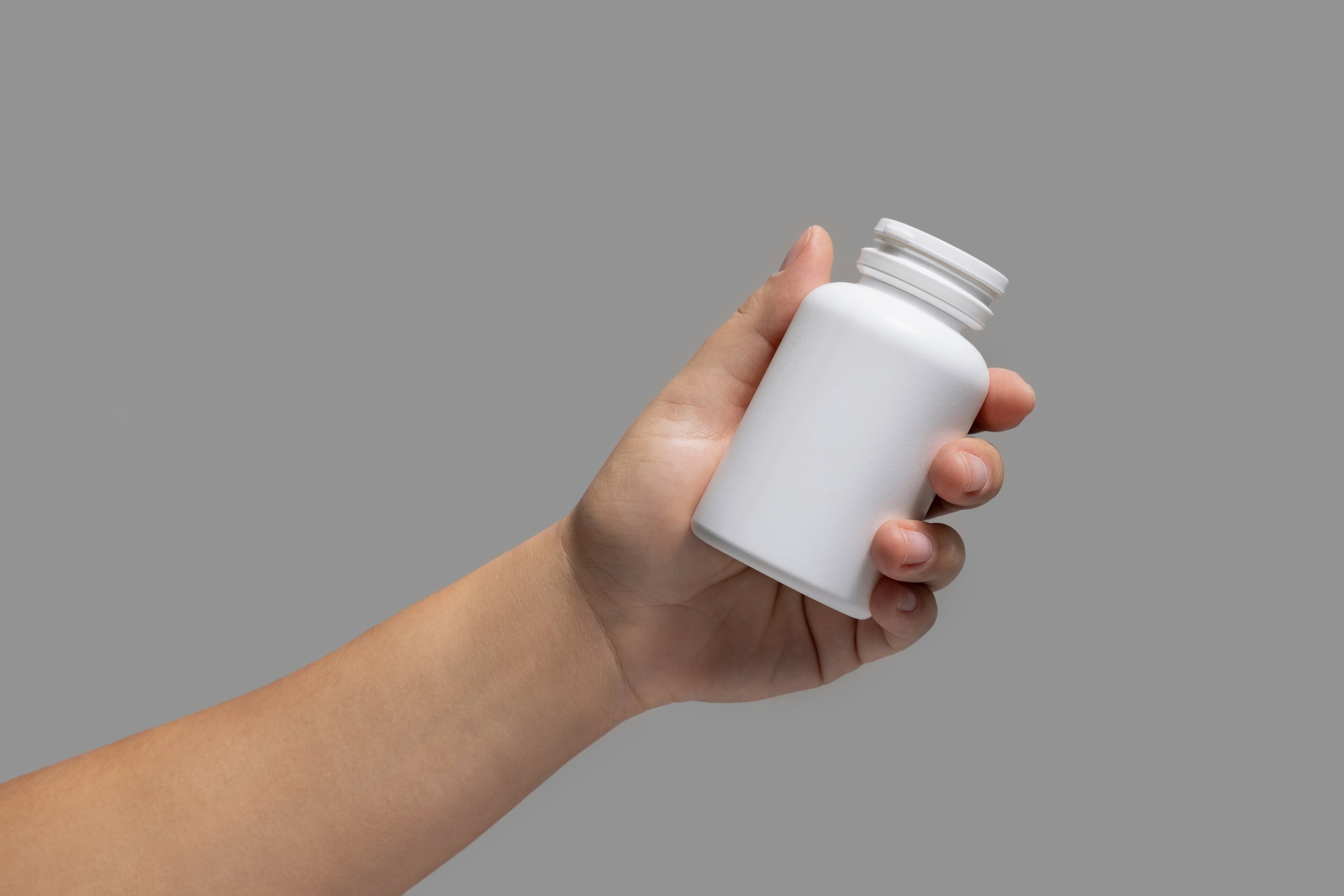 Female hand holding a white plastic pill bottle closeup against grey background.  Packaging mockup for your pills or capsules herbal supplement