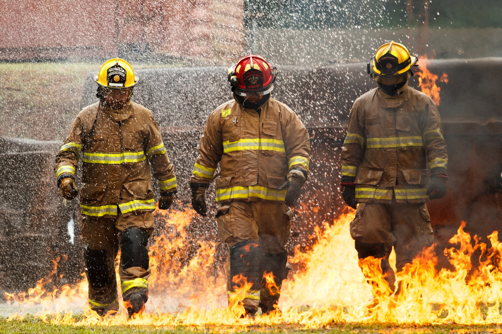 Firefighters in full protective gear walk through intense flames and heavy water spray during live fire training exercise. Dramatic scene highlights teamwork, bravery, emergency response.