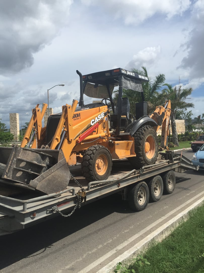 Backhoe loader on a flatbed trailer parked on a road under a cloudy sky.
