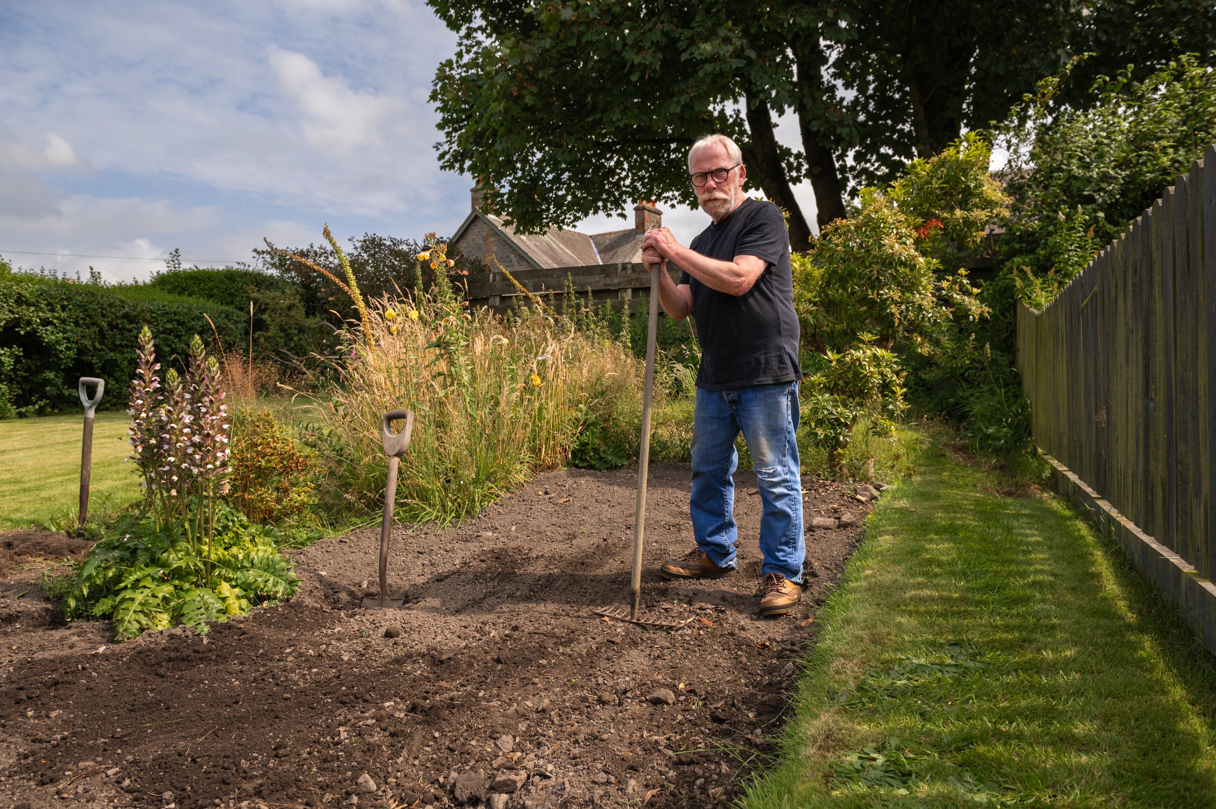 Senior man standing in a flowerbed holding a rake on a summer morning in Scotland