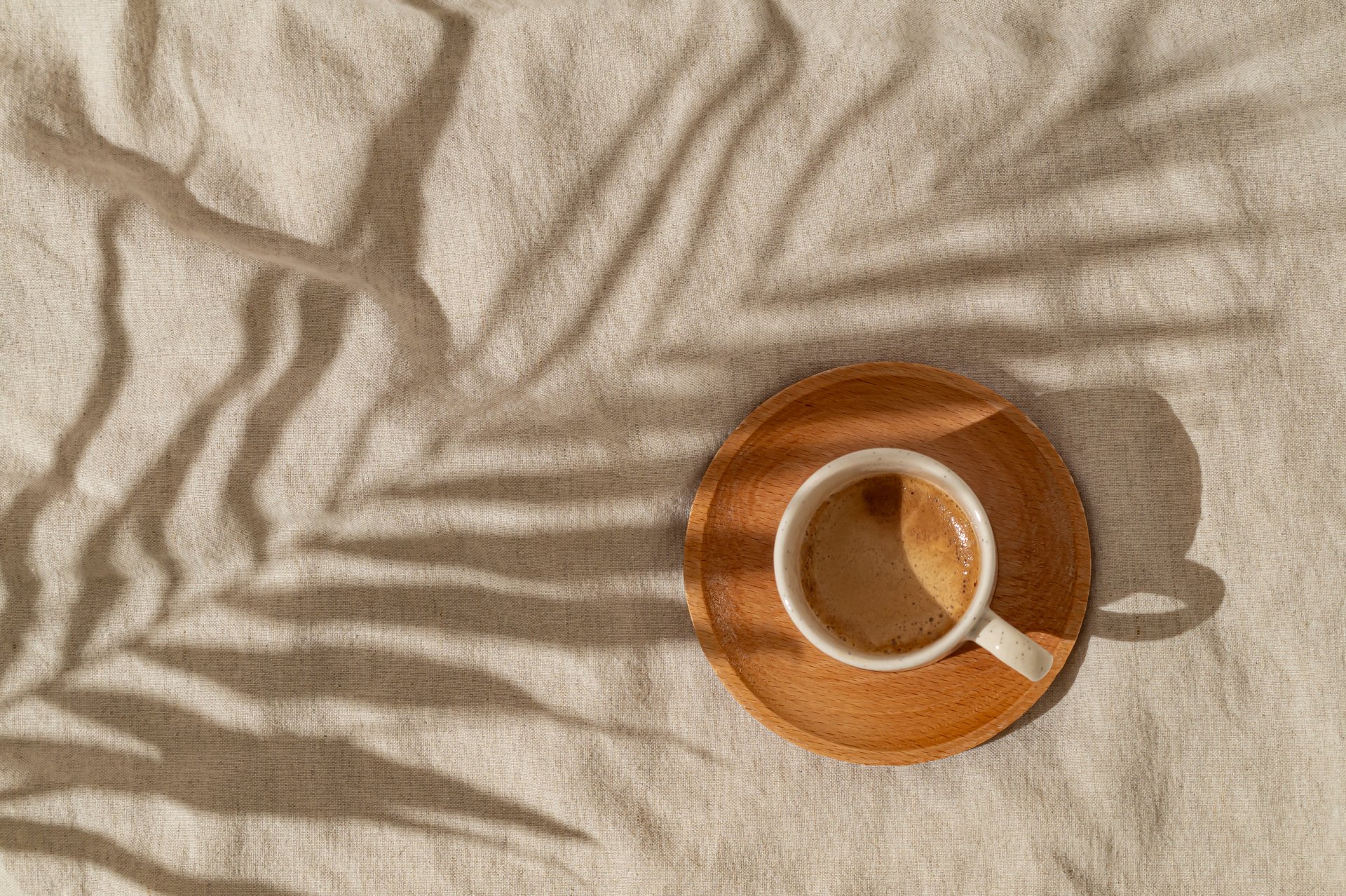 Summer day scene made with cup of coffee and tropical palm tree leaf shadow on sand color linen background. Sun and shadows. Minimal summer coffee concept. Flat lay style. Top view.