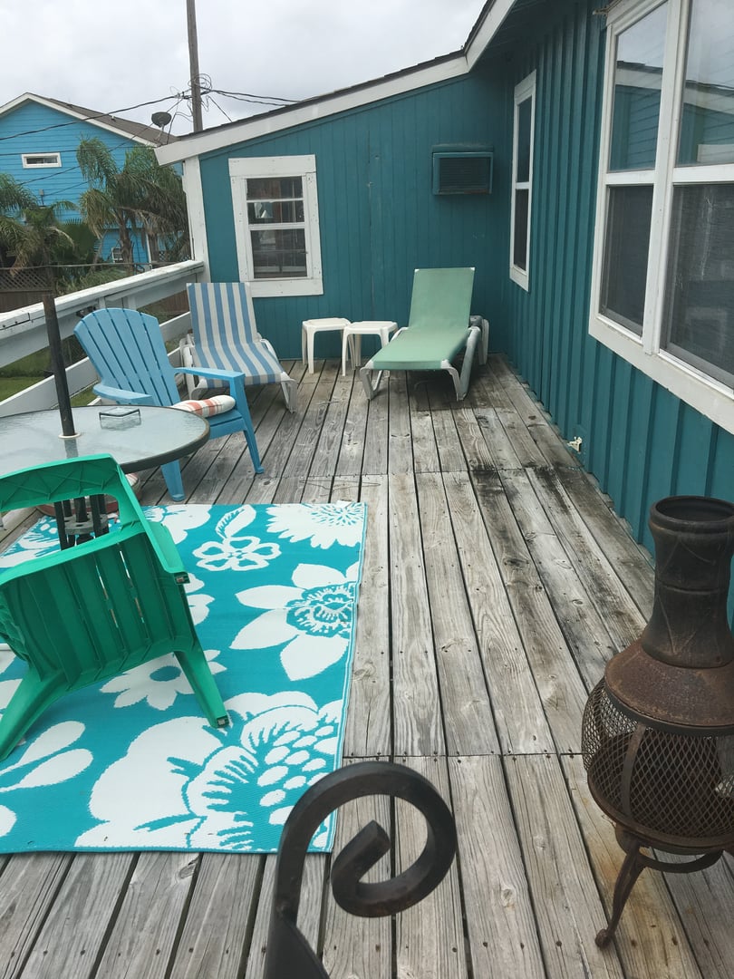 Wooden deck with colorful chairs, a patterned rug, and a chimney. Adjacent houses and palm trees are visible in the background.