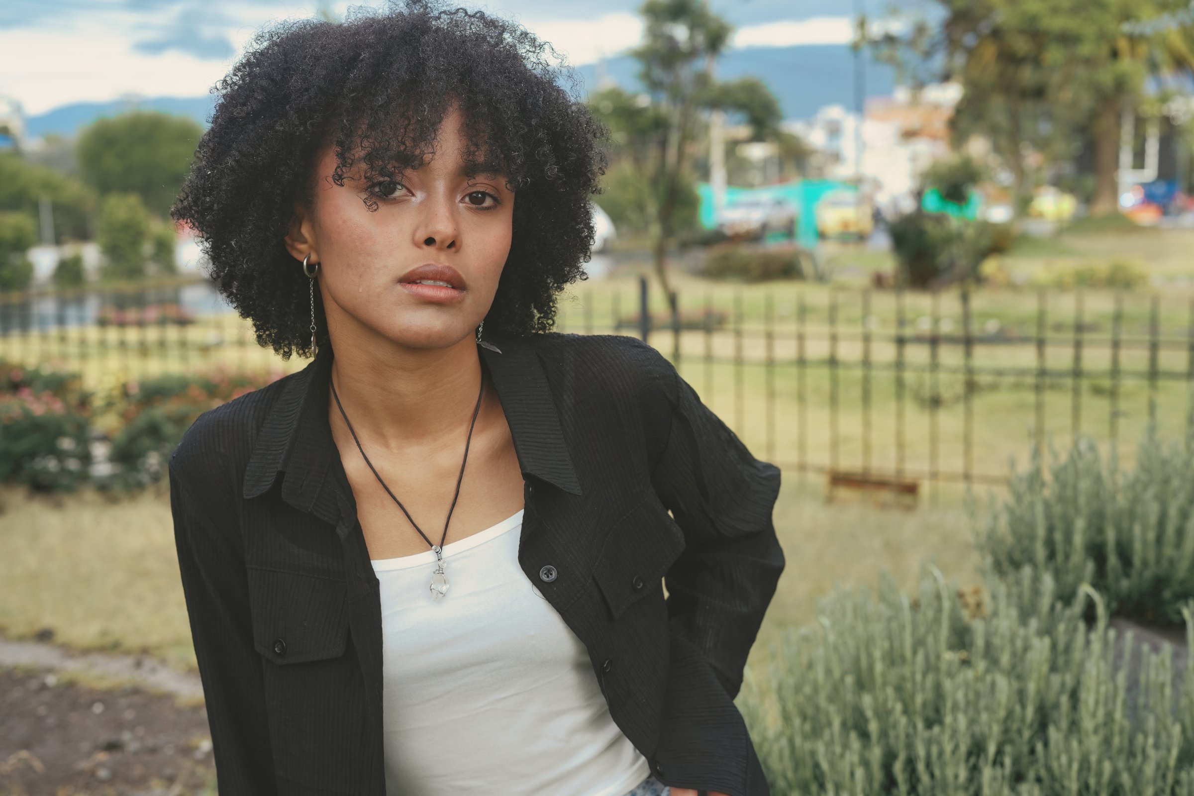 Confident young adult black woman with natural curly hair standing in an urban park, showcasing street style fashion and individuality while looking directly at the camera