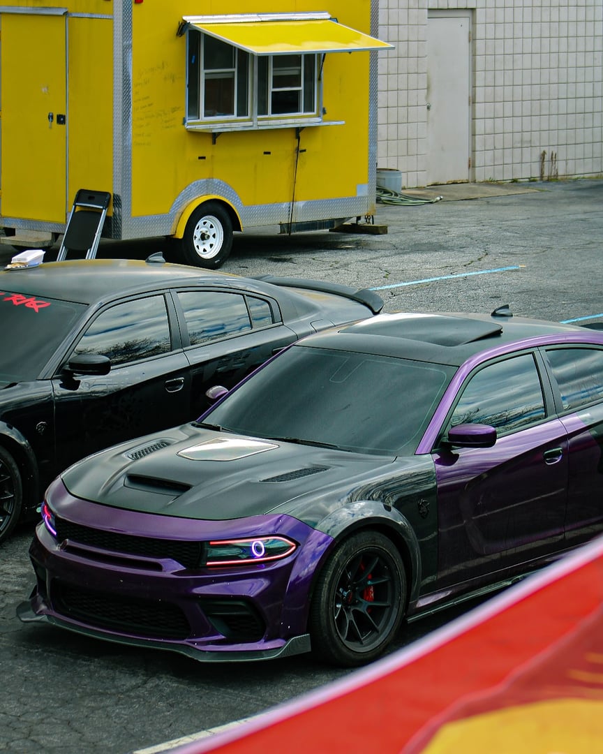 Two Dodge muscle cars, one in black and one in purple, parked near a yellow food truck in a parking lot.