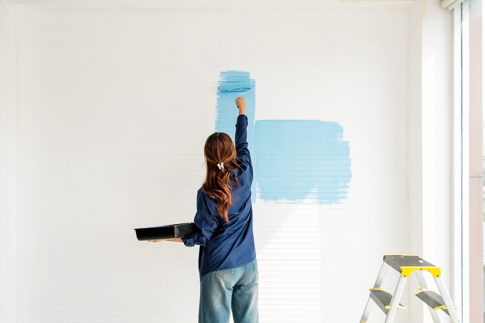 Rear view of a woman painting a white wall with blue paint using a roller during a DIY home renovation project. Bright interior scene captures creativity, self-reliance, and home improvement.