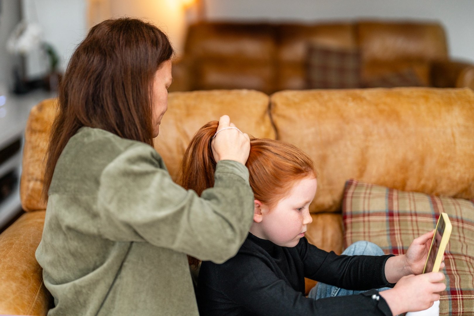 A mother is styling her daughter's hair as the child focuses on a tablet, sitting comfortably on a couch in a warm, inviting living room setting.