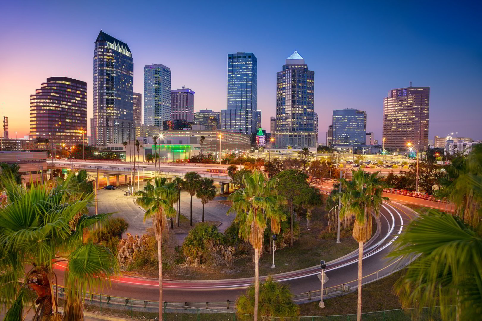 Tampa's skyline features modern high-rise buildings against a twilight sky transitioning from warm orange to deep blue. Palm trees frame the view, with illuminated streetlights and streaks of car lights indicating motion. Prominent architecture includes glass and concrete towers, reflecting the city's urban stature. The vibrant mix of light and shadow captures Tampa's lively atmosphere as day turns to night.