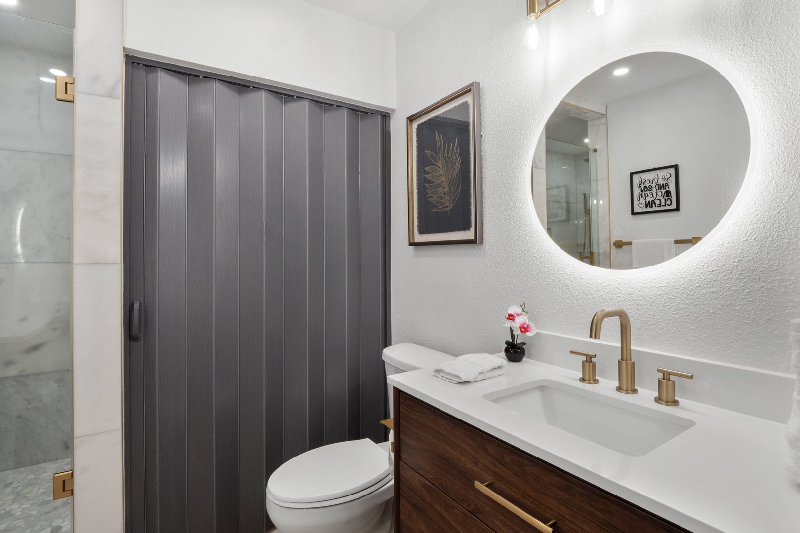 Modern bathroom with circular mirror, wooden vanity, gold fixtures, and sliding door beside the glass shower.