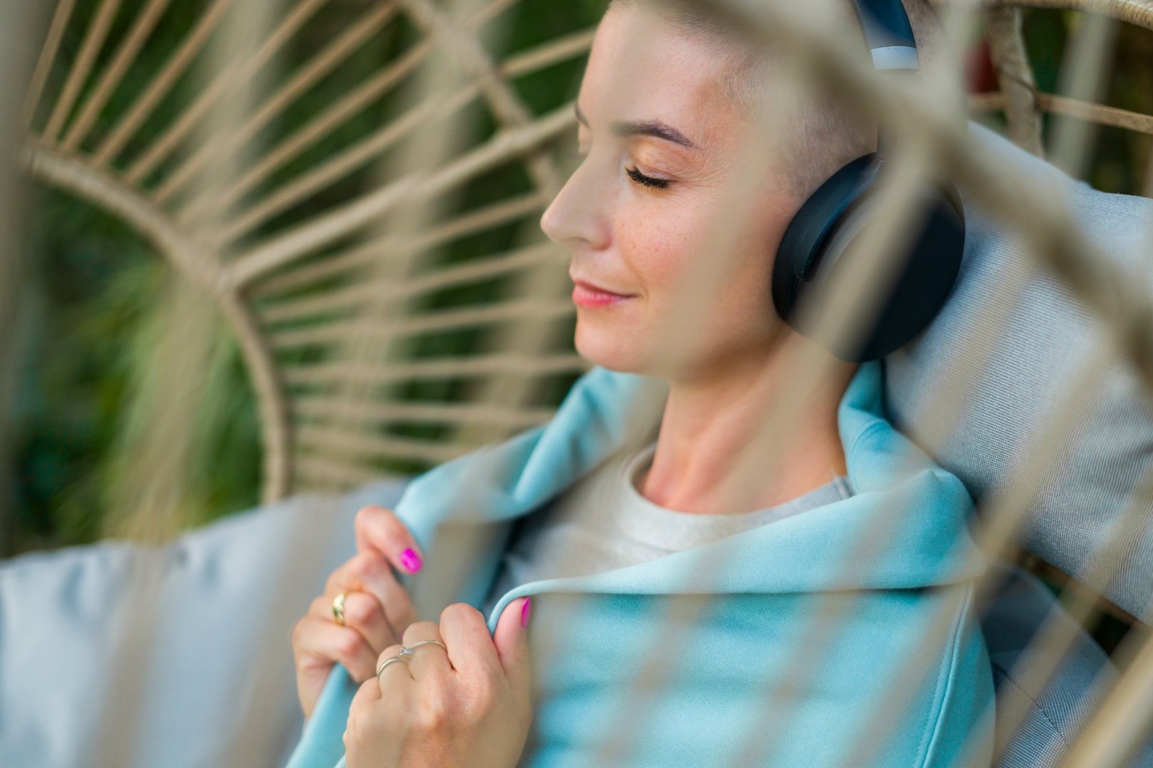 Young female cancer patient relaxing in hanging chair on the garden patio, listening to music. Mental wellbeing during illness.