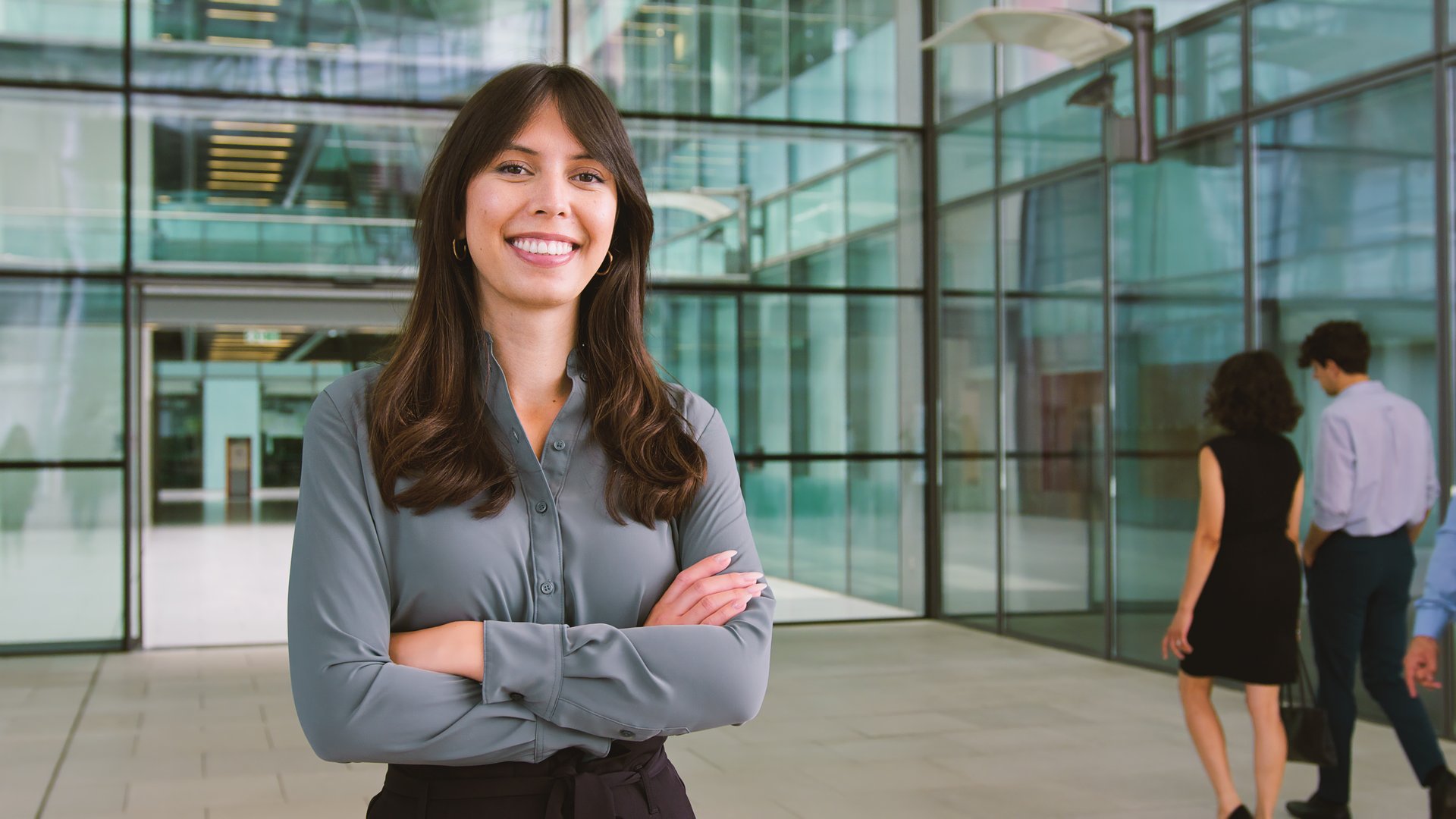 Portrait Of Young Smiling Businesswoman Standing In Lobby Of Busy Modern Office Holding Bag