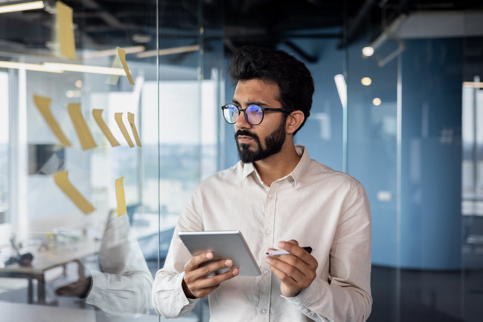 Serious and focused young Indian man working in office with tablet, standing near glass board with notes, processing data, thinking about project.