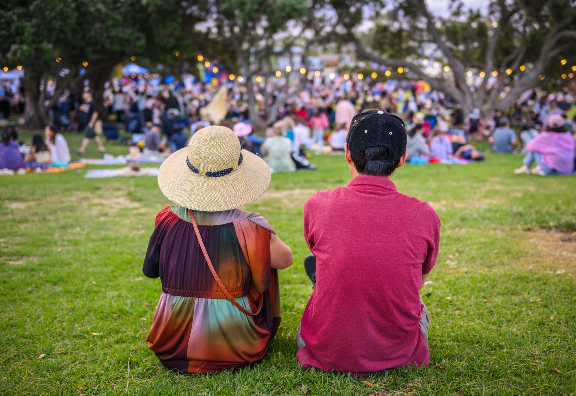 Couple sitting on the grass, unrecognizable crowds listening to the music concert in the park. Auckland.