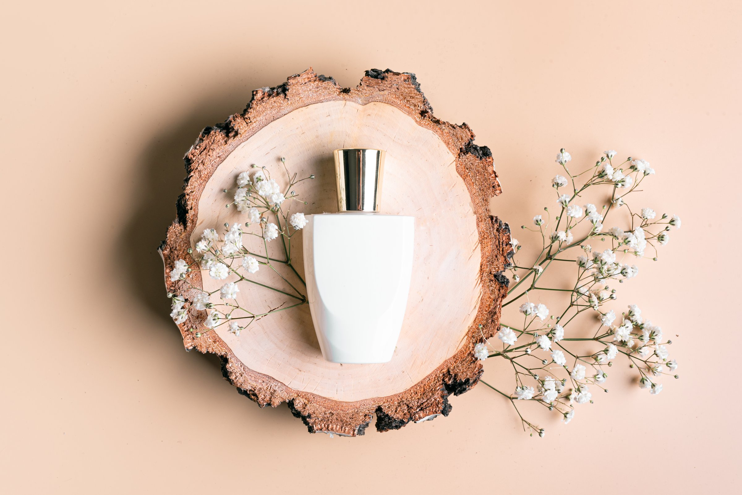 White perfume bottle and gypsophila flowers on wood cut over neutral beige background, top view composition. Perfumery concept. Fragrance branding template.