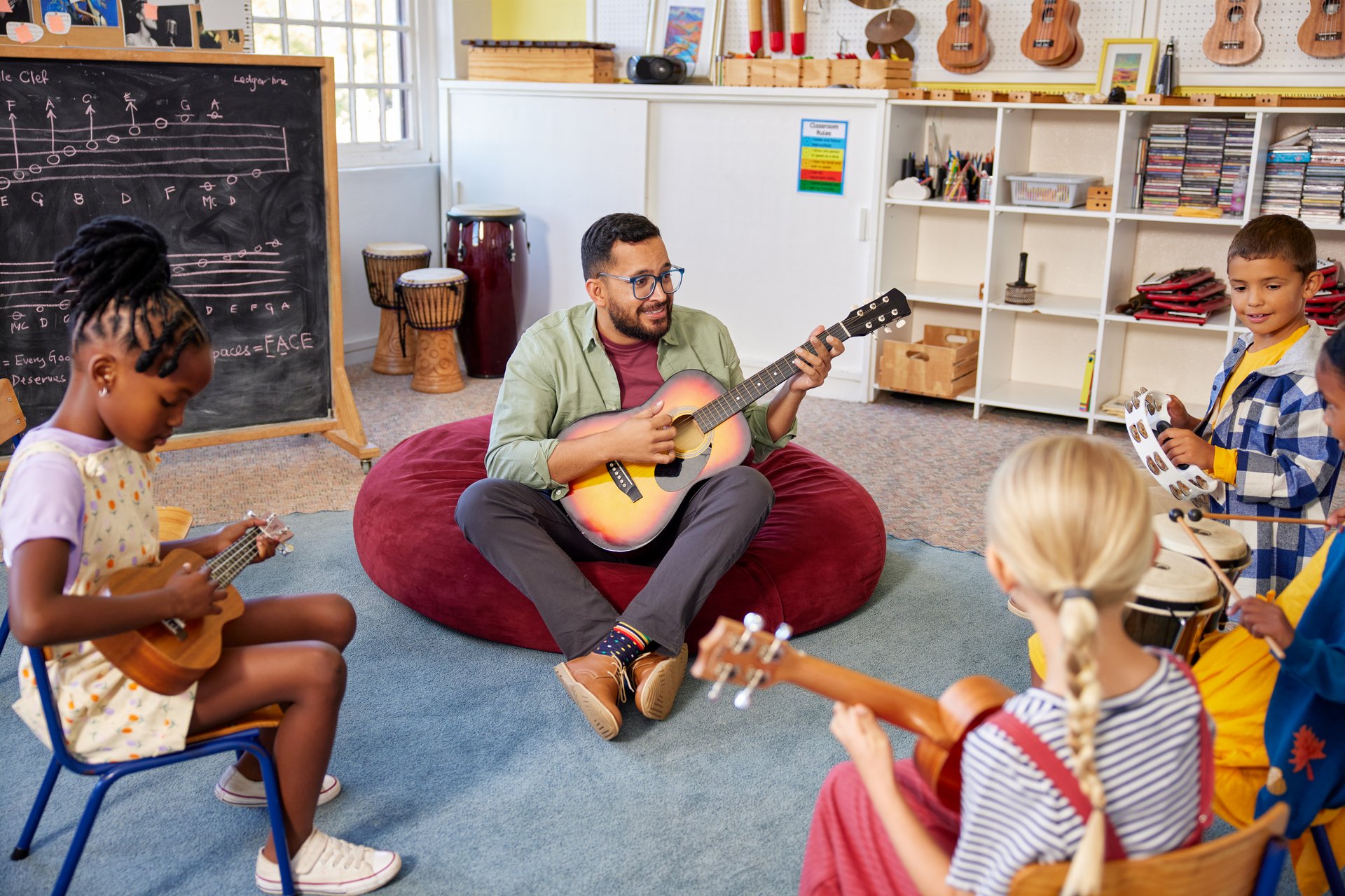 Group of children and teacher playing musical instruments during music class in school. Male teacher playing guitar with pupils while having music lesson in classroom. Professor assisting school children to play musical instruments all sitting in a circle.