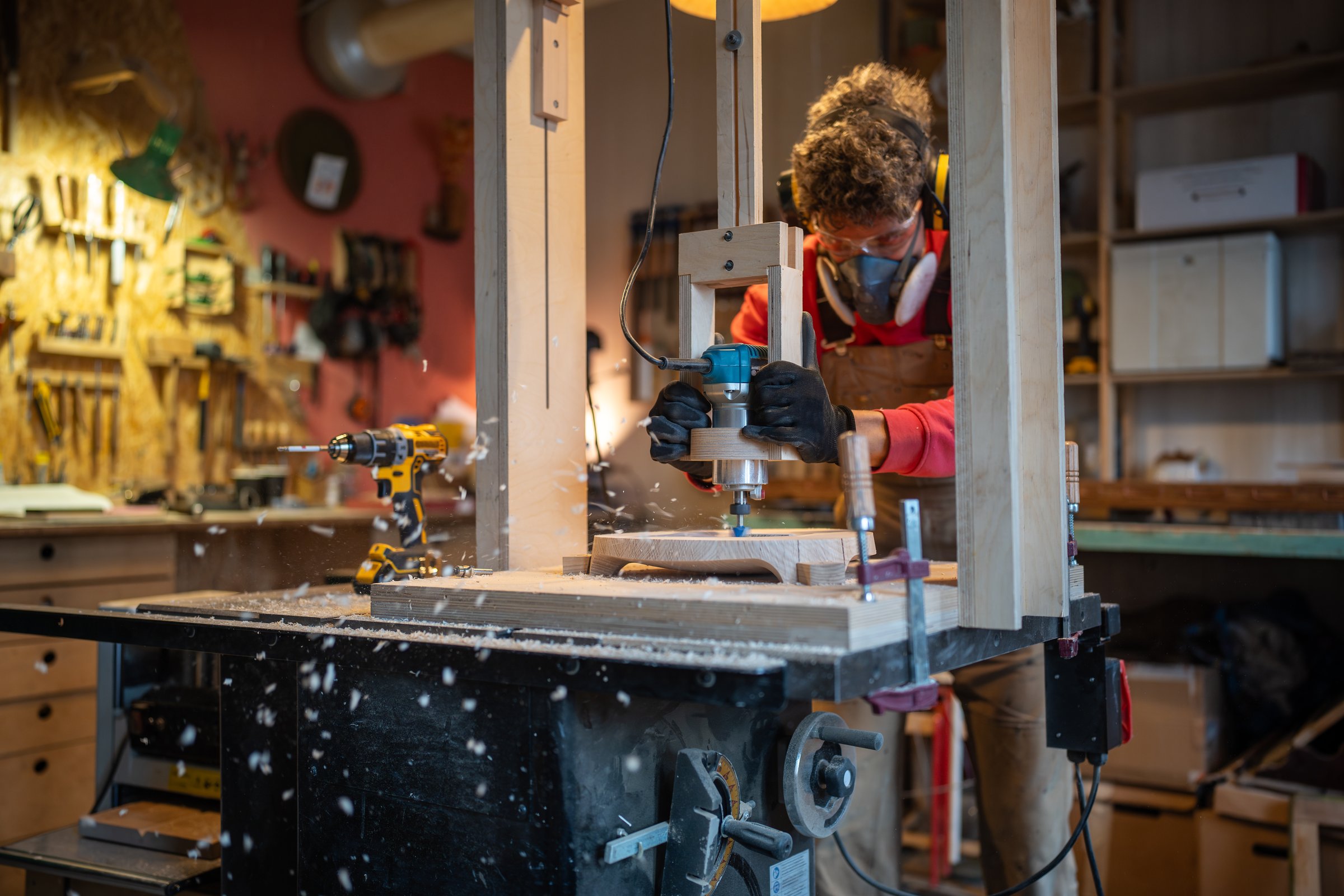 Focused male carpenter wearing protective noise-cancelling headphones, safety googles and respirator milling wooden plank while working in joinery, sawdust is flying around. Woodwork.