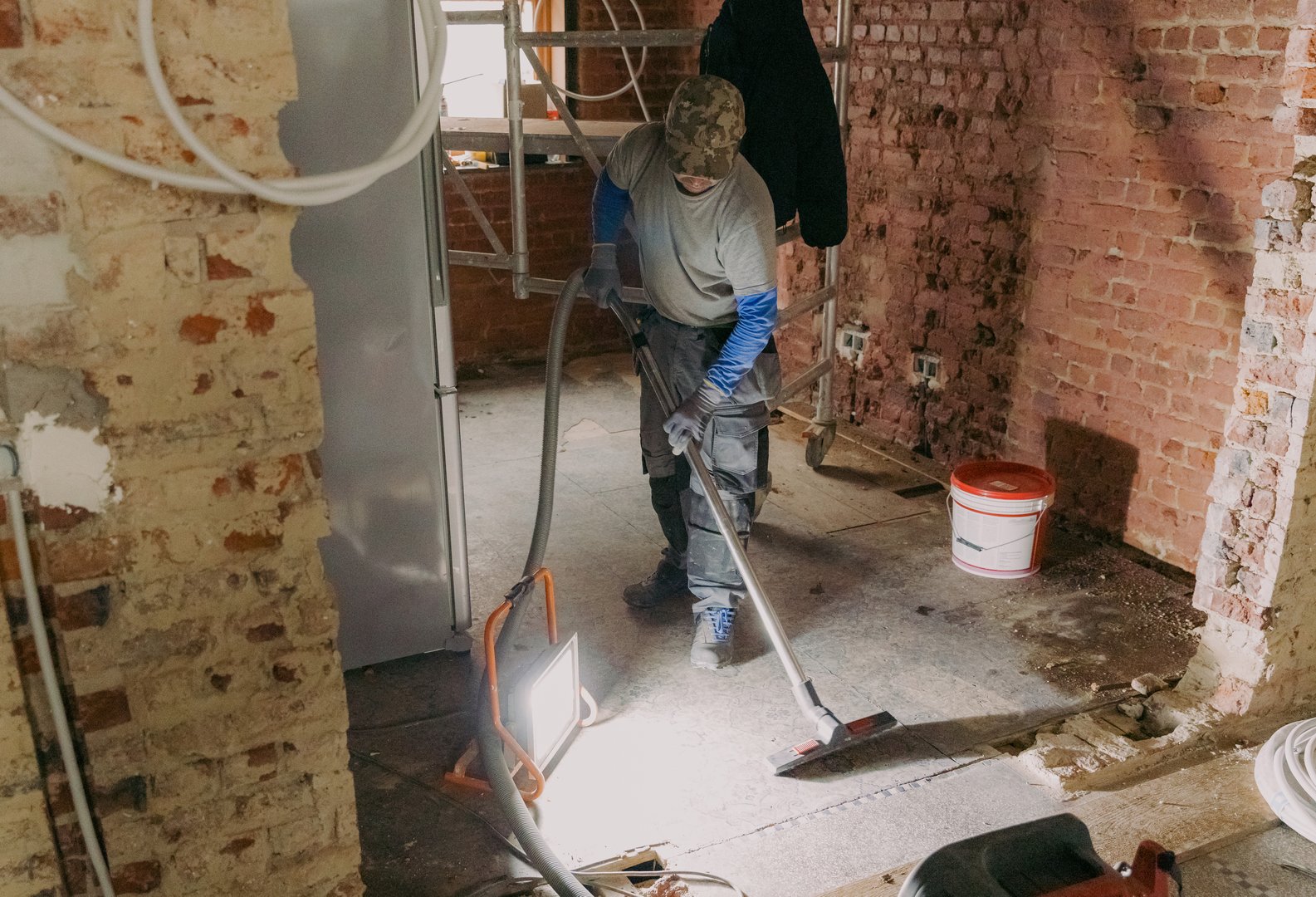 One young Caucasian man in a gray casual work uniform vacuums up construction debris after laying bricks in a room where renovations are underway, standing under the light of a lantern in an old ruined house, close-up side view.