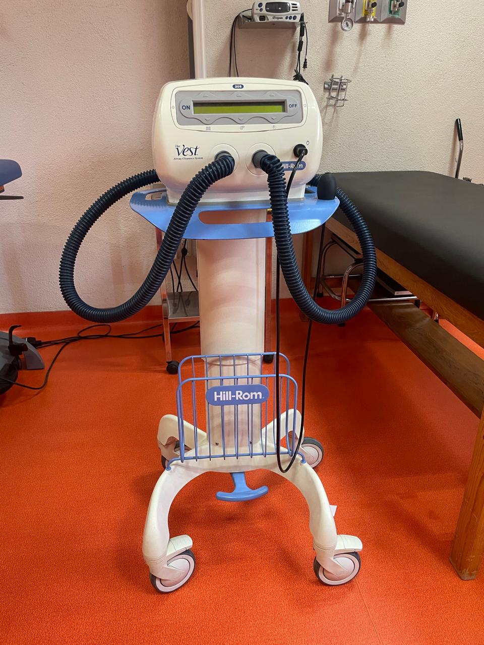 Medical device on wheels with two coiled tubes, labeled "The Vest," in a room with orange flooring and exam table.