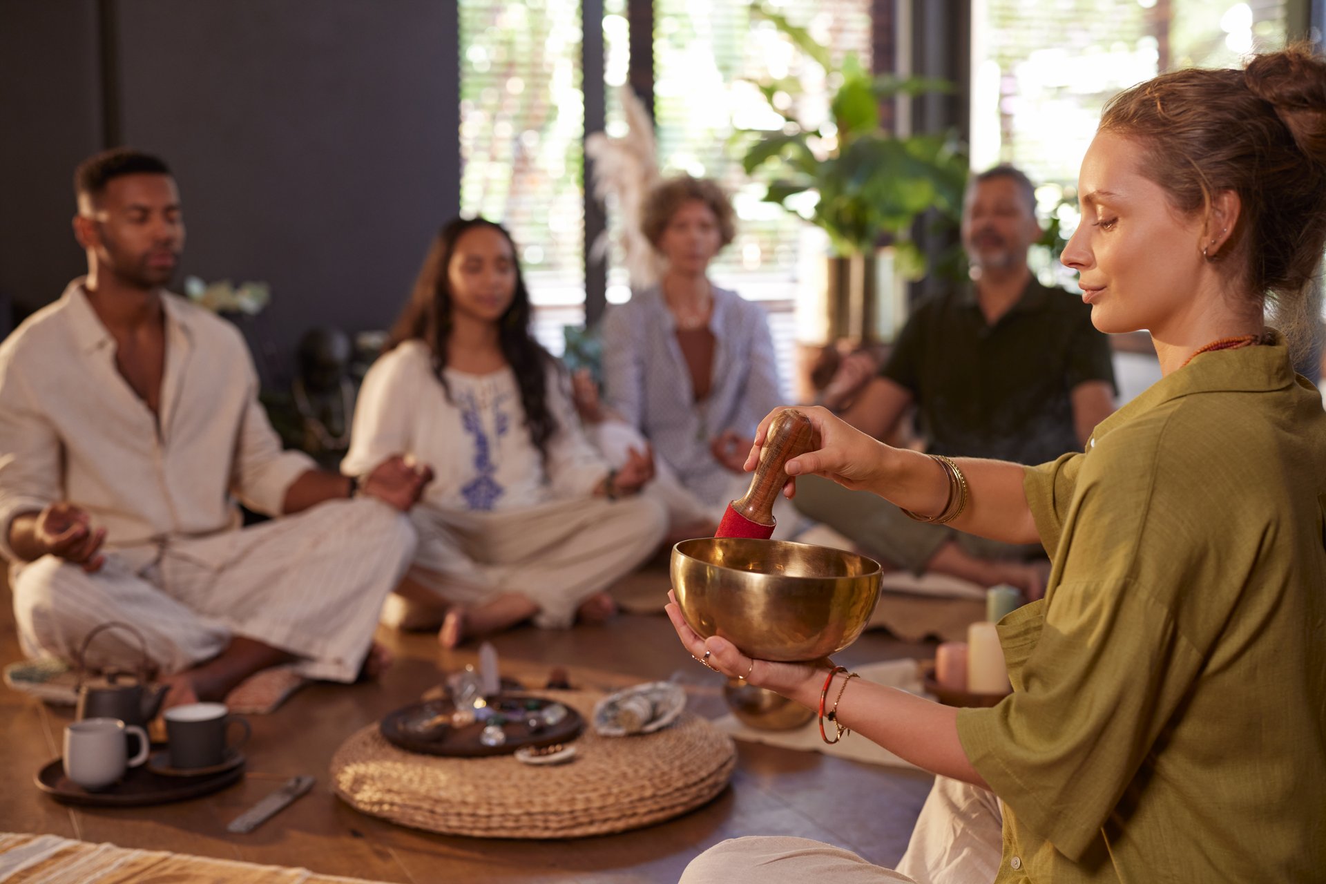 Young woman using singing bowl during healing session at wellness retreat center. Group of multiethnic people meditating in background, practicing mindfulness and inner peace. Tibetan singing tibetan bells in sound therapy at wellness center, energetic cleansing concept.