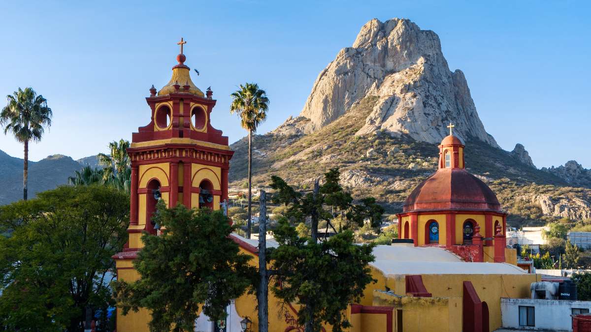 A picturesque church with red and yellow domes, set against a backdrop of a large rocky hill and palm trees under a clear sky.