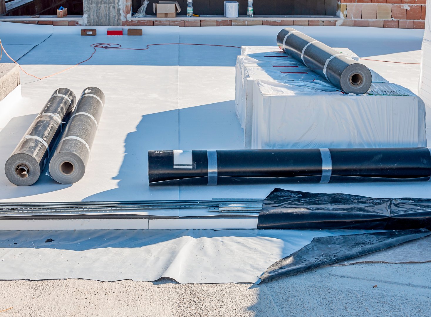 Construction site with a flat roof being waterproofed. The image shows a partially installed waterproof membrane, tools, and construction materials. The scene highlights the roofing process.