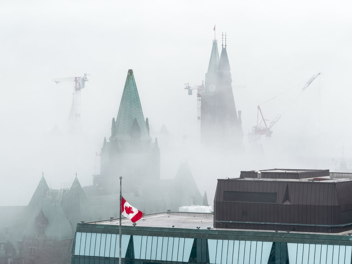 Moody foggy morning at Parliament Hill in Ottawa, Ontario, Canada, with Peace Tower partially obscured by mist, Canadian flag waves in the foreground, while cranes loom in the background (April 2025)