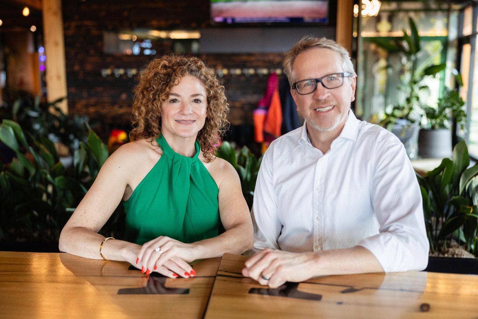 A woman in a green dress and a man in a white shirt smiling, seated at a wooden table in a lush indoor setting.