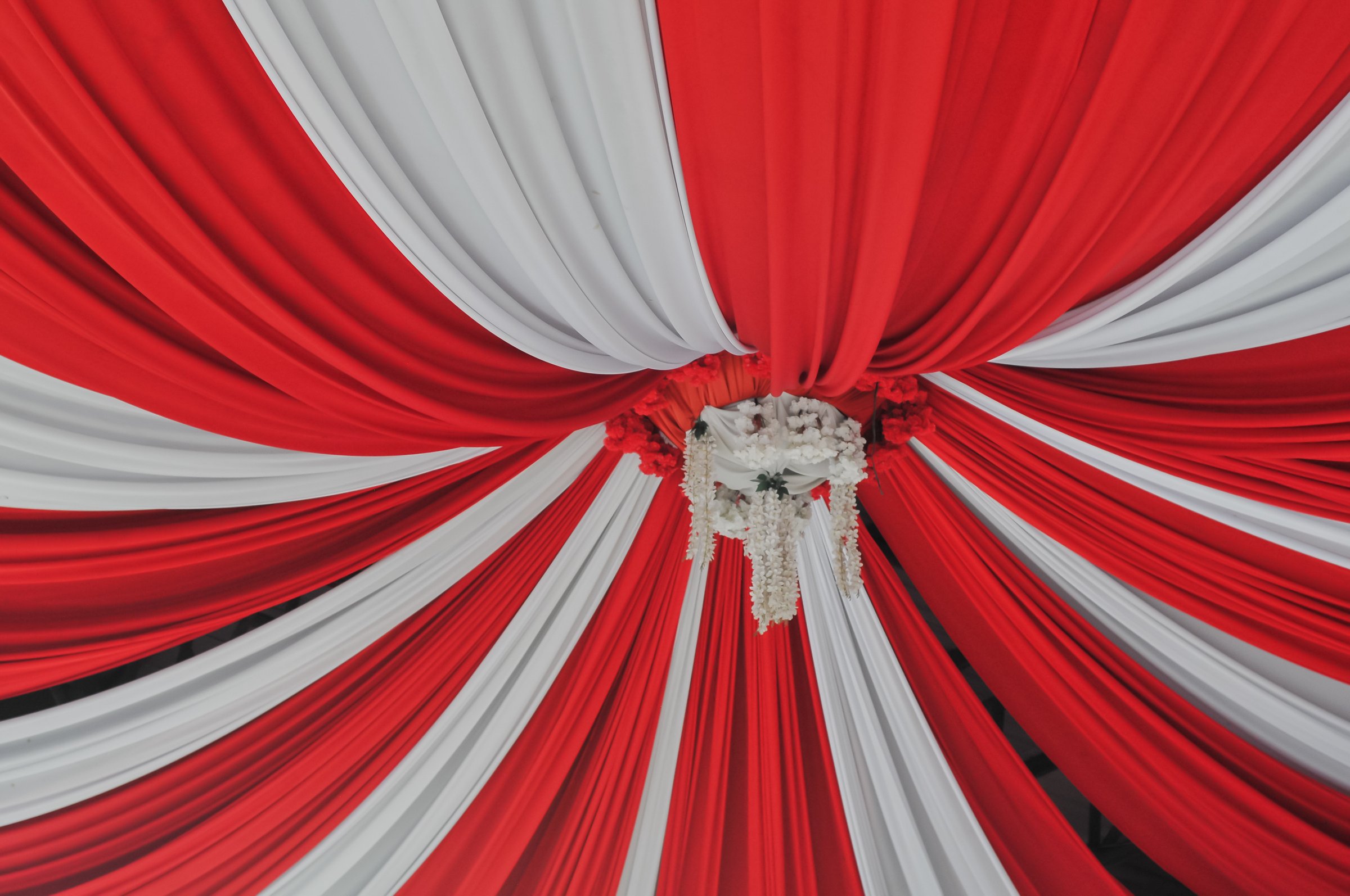 A visually striking pattern of alternating red and white draped fabric radiates from a central white floral chandelier, forming a canopy ceiling, symbolizing national colors, festivity, and wedding or