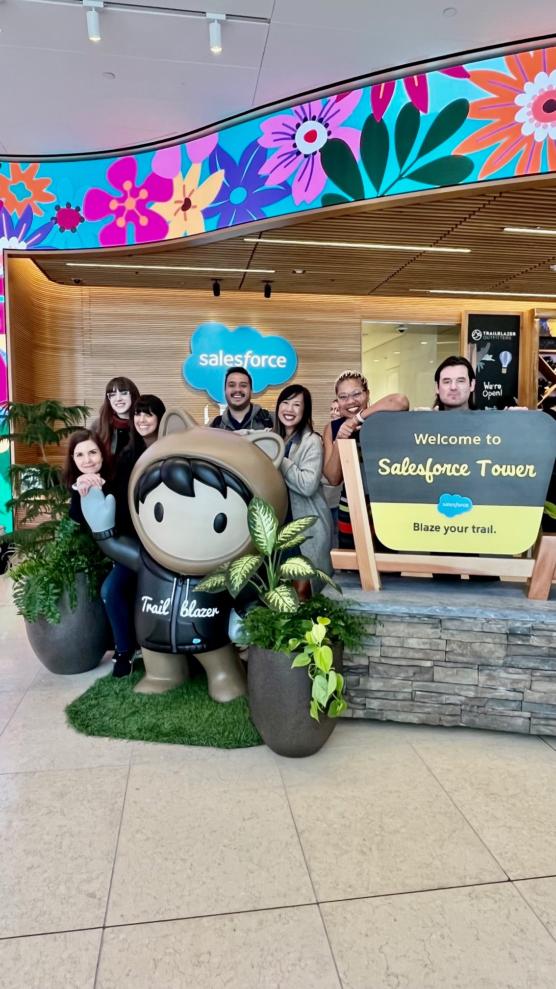 Group of people posing with a Salesforce mascot in front of a "Welcome to Salesforce Tower" sign and colorful mural.