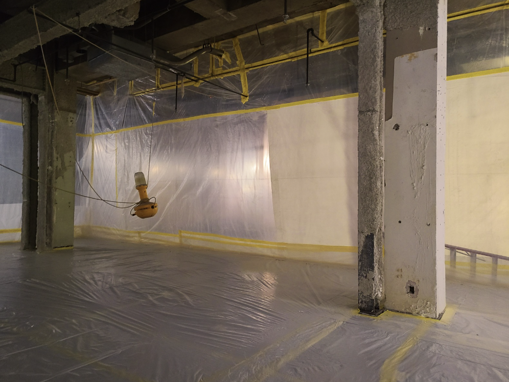 Construction site interior with plastic sheeting covering walls and floor, suspended light fixture, and exposed beams.