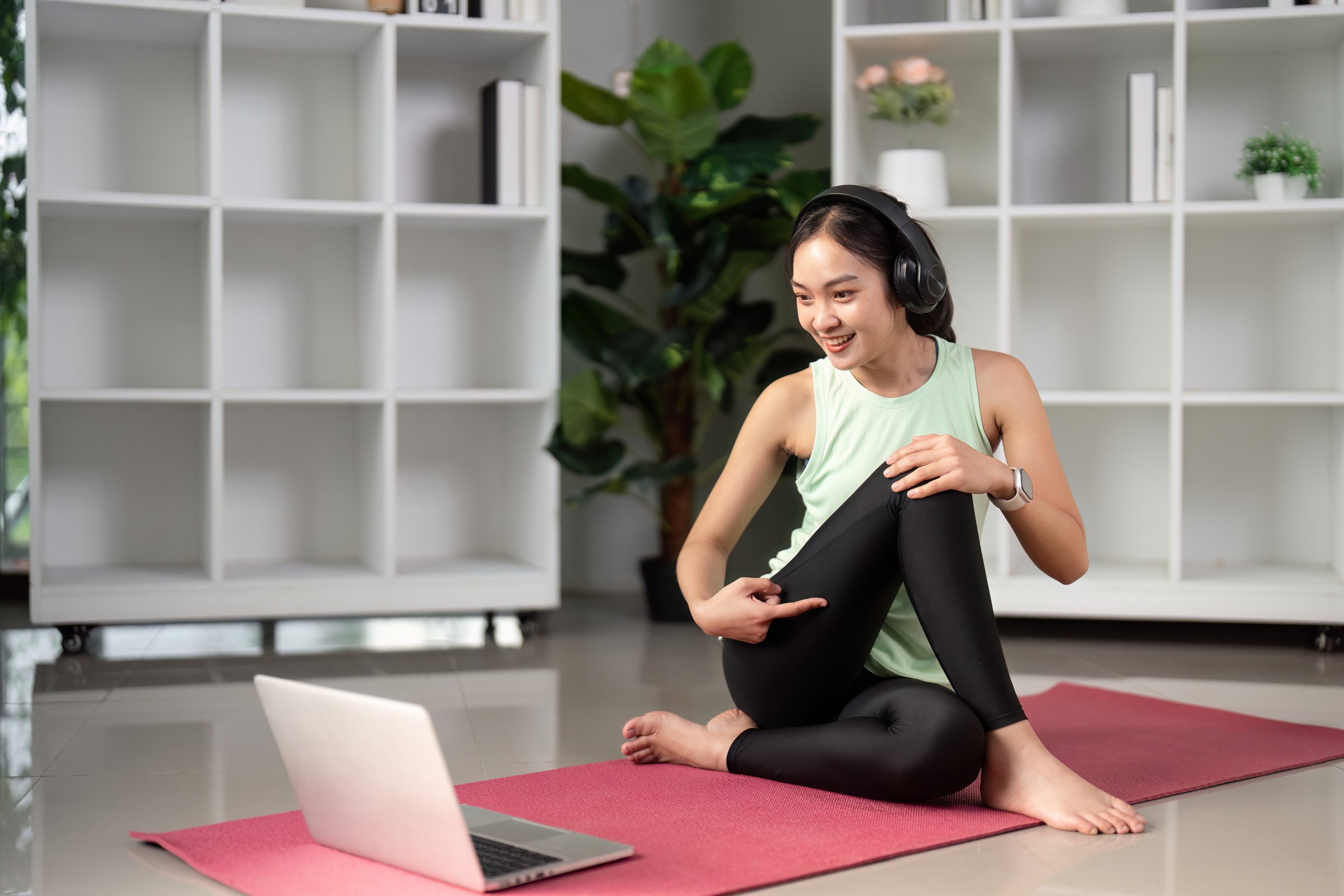 A woman sits on a yoga mat, engaged in an online fitness session, combining technology with wellness practices.