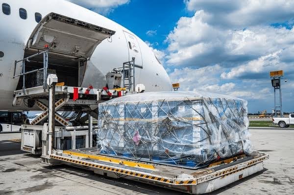 An aircraft being loaded with a large, wrapped cargo pallet on a sunny day at an airport.